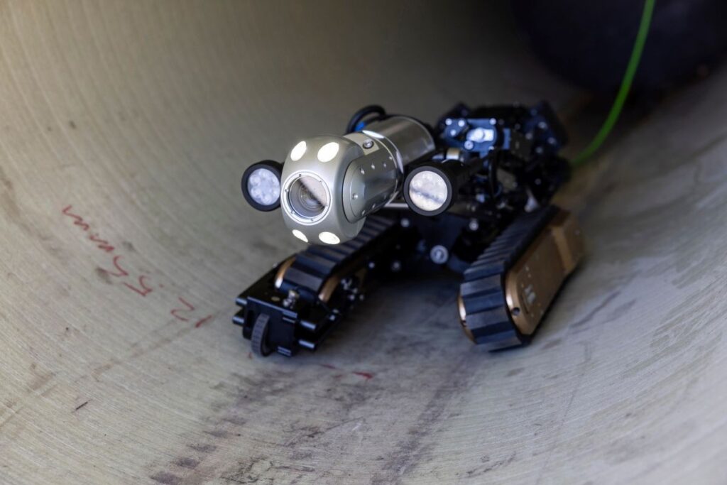 Tracked robotic inspection crawler equipped with high-intensity lights and a camera, performing a visual inspection inside an industrial pipe.